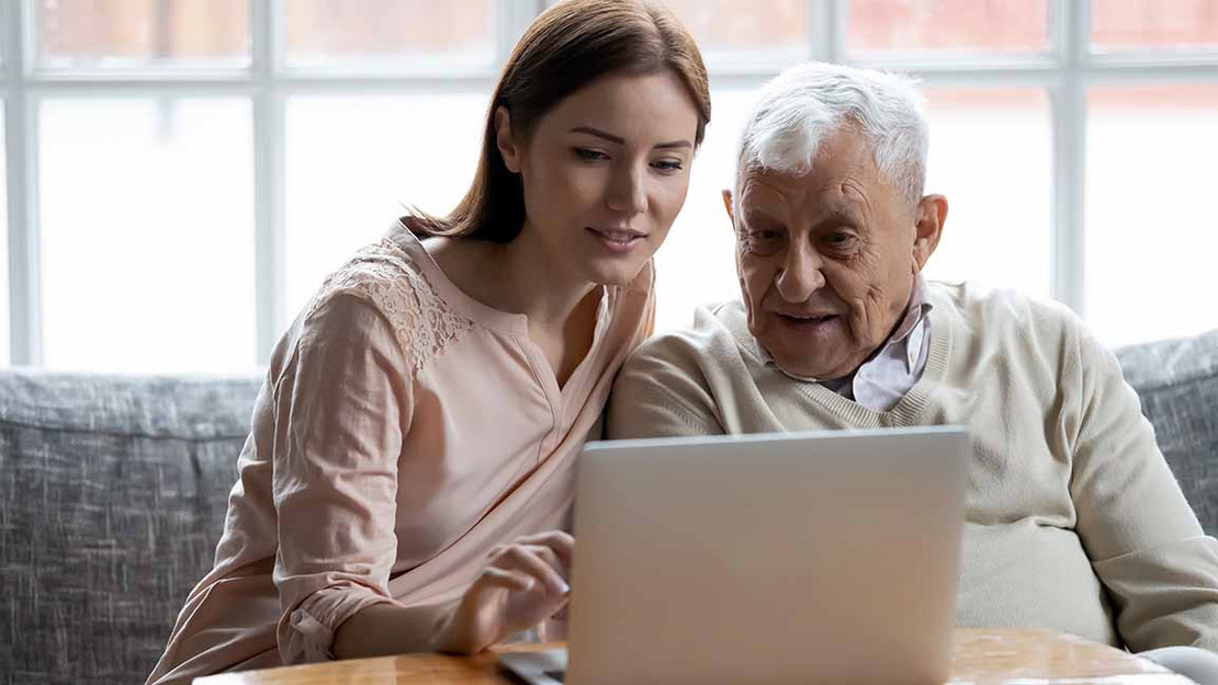 Jüngere Frau sitzt mit altem Mann vor einem Laptop