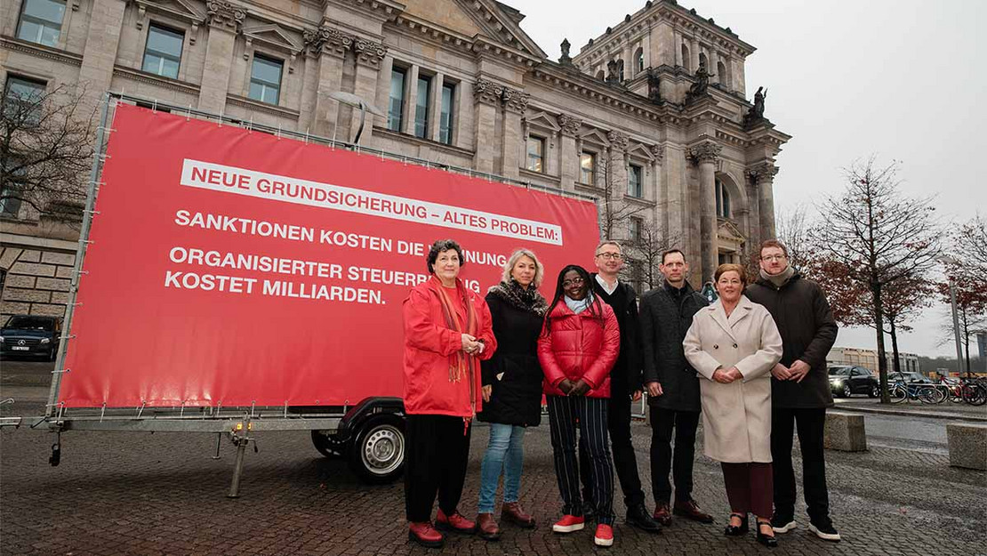 Sieben Personen am Bundestag vor einem Plakat mit der Aufschrift: Neue Grundsicherung – altes Problem: Sanktionen kosten die Wohnung. Organisierter Steuerbetrug kostet Milliarden.“