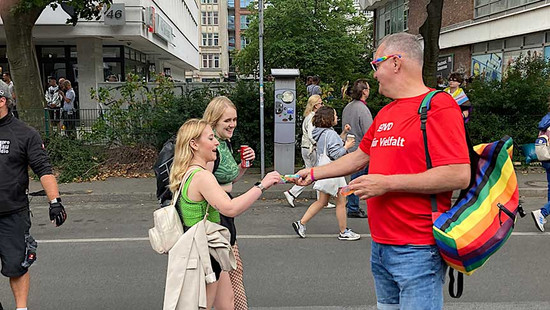 Mann im roten SoVD-Shirt gibt zwei jungen Frauen SoVD-Bändchen. 