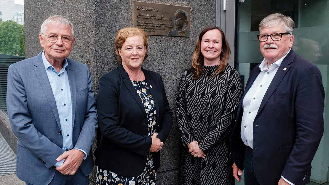 Vier Personen vor einer goldenen Plakette an einer Hauswand. 