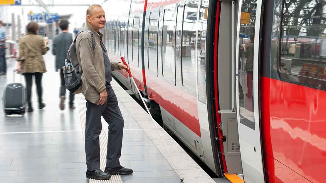 Blinder Mann steht auf einem Bahnhof an einem Zug mit offenen Tür. 