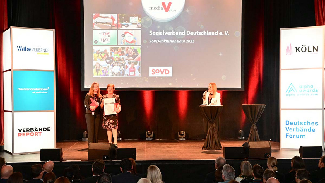 Zwei Frauen stehen auf einer Bühne, im Hintergrund eine LED-Leinwand. Rechts eine Moderatorin am Pult. 