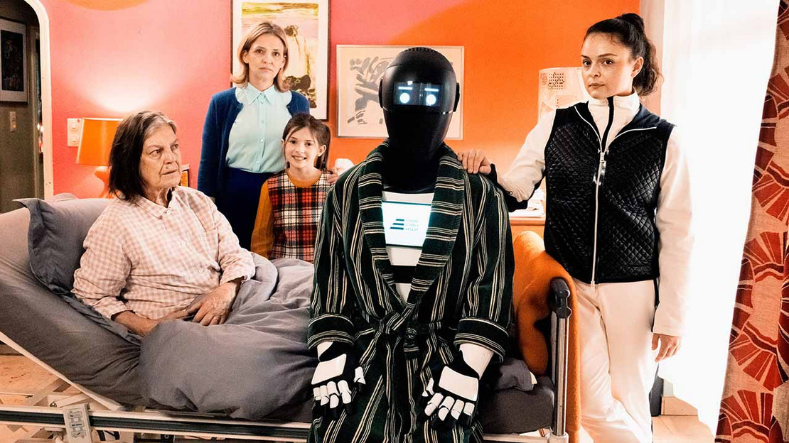 Filmszene aus „Frau Winkler verlässt das Haus“. Ein Pflegeroboter sitzt neben einer älteren Frau auf dem Bett, im Hintergrund drei weitere Personen. 
