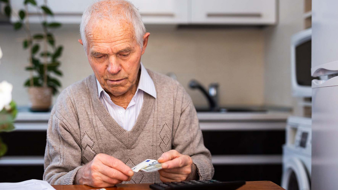 Älterer Mann sitzt mit Taschenrechner und Geldschein in der Küche.