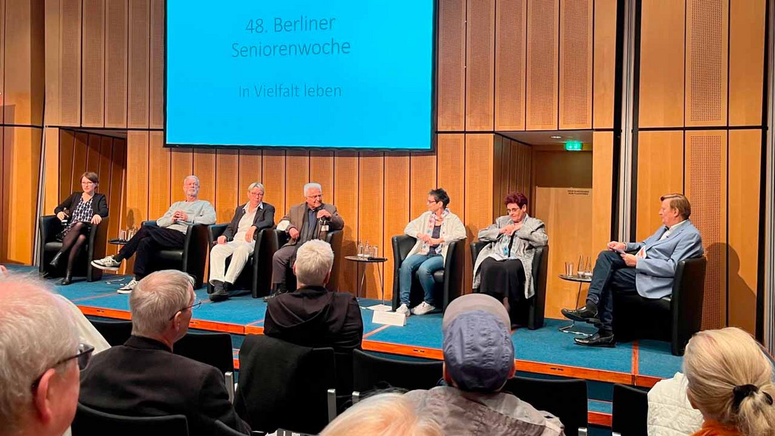 Blick in Veranstaltungsraum. Sieben Menschen sitzen auf der Bühne. 