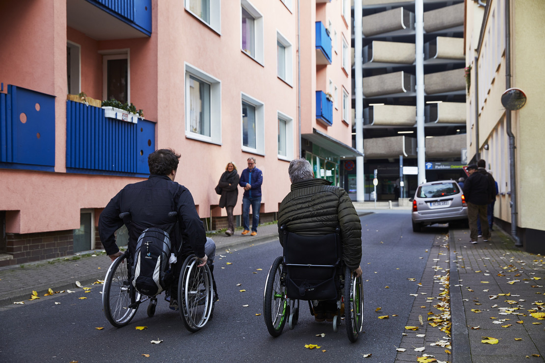 Zwei Menschen im Rollstuhl fahren durch eine Straße. 
