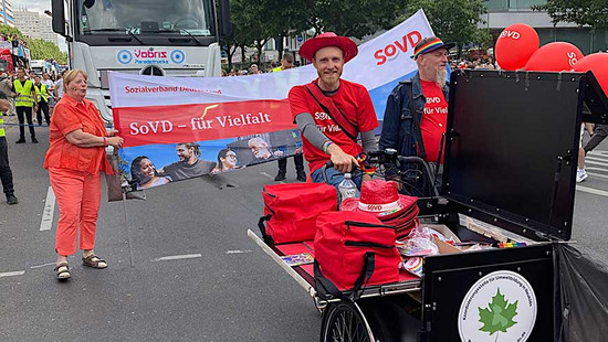 Mann fährt ein Lastenrad. Frau dahinter hält ein Banner mit der Aufschrift "SoVD für Vielfalt"