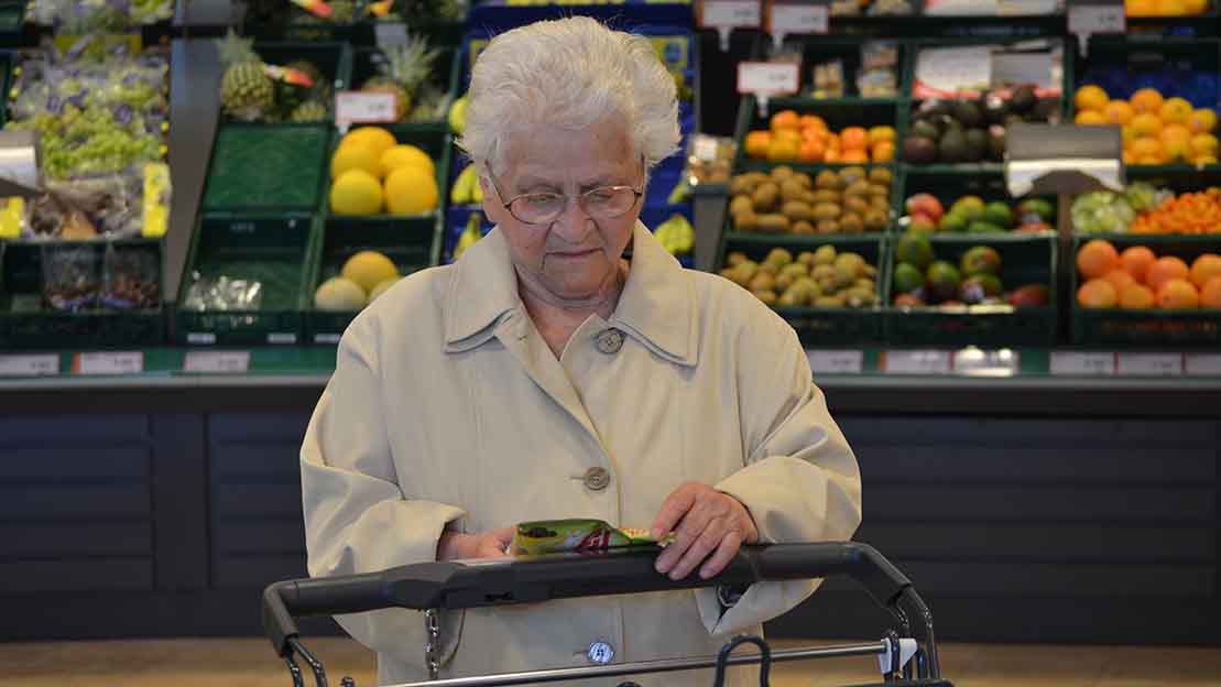 Alte Frau mit Einkaufswagen vor dem Obstregal in einem Supermarkt. 